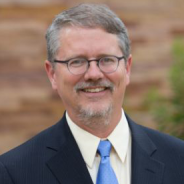 White male wearing a blue suit with grey hair and glasses.